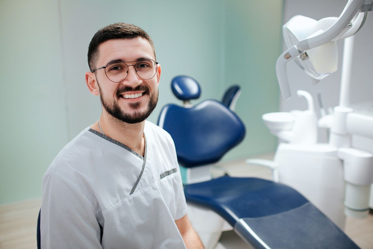 Positive young bearded dentist sit in dentistry room and pose on camera. He wear white robe and glasses.